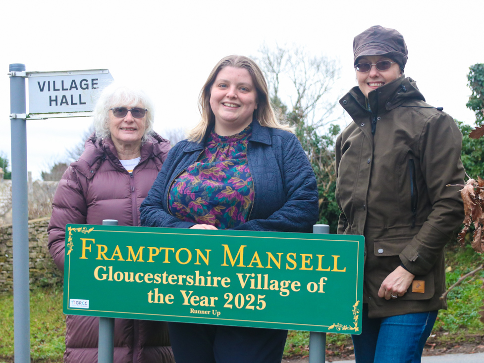 Parish Clerk Babs Maloney, GRCC's Sabrina Dixon, and village volunteer Lucy Cameron-Davies stand behind a sign which reads Frampton Mansell, Gloucestershire Village of the Year 2025, Runner Up (with the last two words in a very small font)