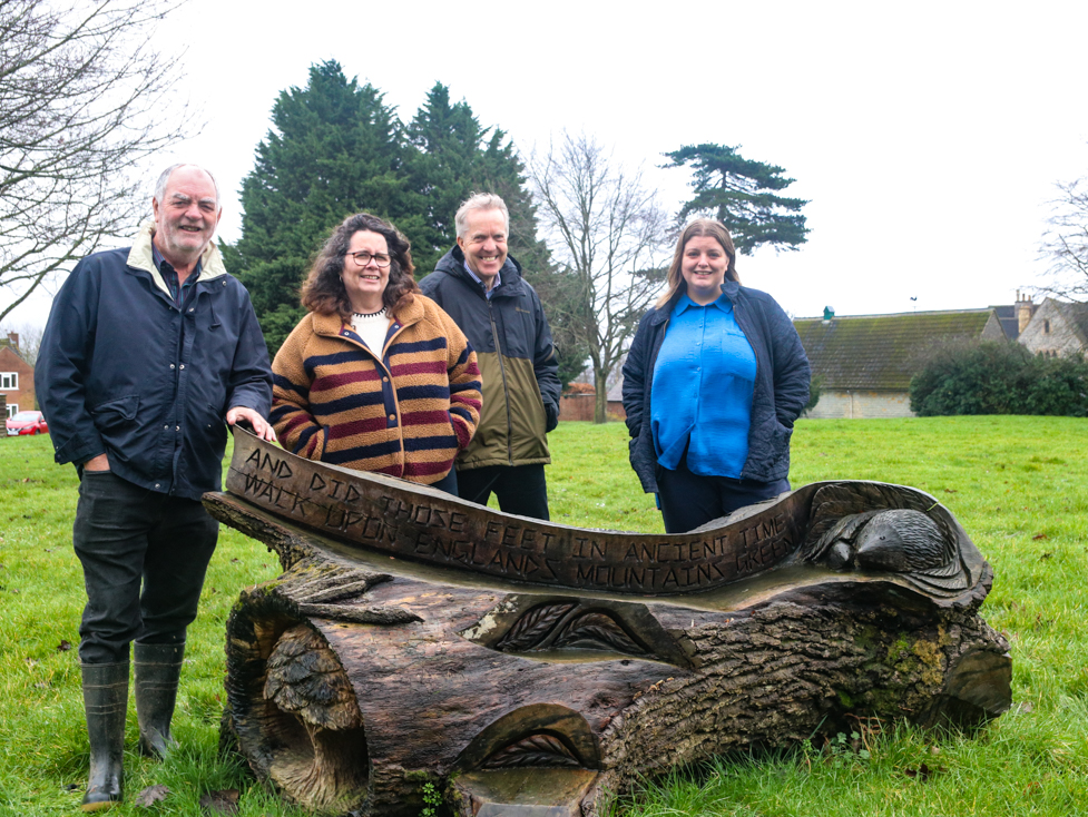 People standing behind a bench carved from a treetrunk featuring animals and leaves,  on a green space in Highnam