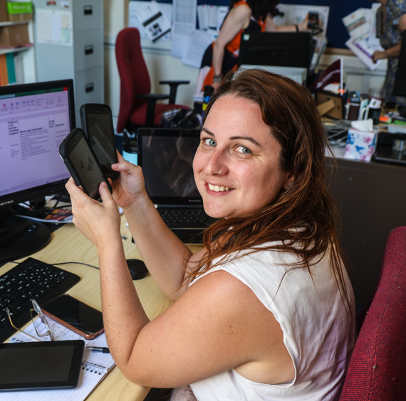 Photo of our link worker smiling and holding two mobile phones sitting at a desk with lots of computer monitors